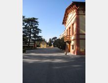 Cortile o viale asfaltato in un complesso con edifici bassi; a destra una palazzina color crema e rosso con balconcino in legno e tetto sporgente. L’area è circondata da alberi e piante in vaso sotto un cielo sereno.