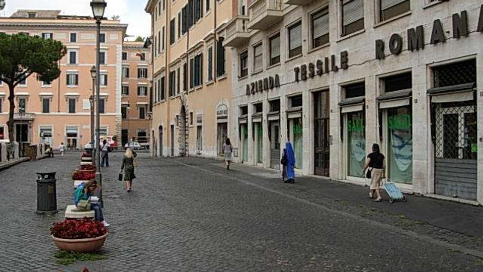 Strada urbana lastricata con edifici storici in stile italiano, con vetrine al piano terra e l’insegna “Azienda Tessile Romana” sulla facciata. Sul lato sinistro si vedono lampioni, fioriere e un grande albero, con la via che curva tra i palazzi sullo sfondo.