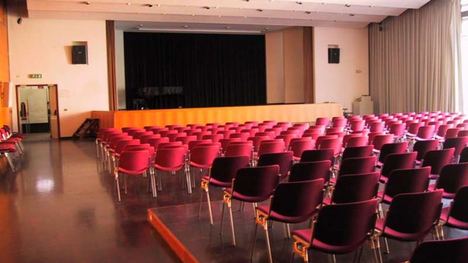 Sala auditorium con file di sedie imbottite rosse e nere disposte verso un palco rialzato con sipario nero. Soffitto con faretti incassati, pavimento lucido e grandi finestre laterali con tende che illuminano l’ambiente.