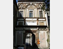 Facciata di un edificio storico con decorazioni in stucco e medaglioni, finestre alte incorniciate e una grande arcata centrale che funge da passaggio verso una strada stretta. Alla base si vede un portale ad arco in pietra con bassorilievi e cartelli stradali accanto, in un contesto urbano tra edifici ravvicinati.