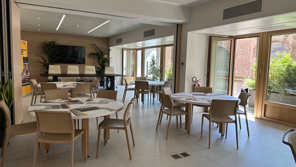 Sala riunioni moderna con tavoli rotondi e sedie beige su pavimento chiaro, illuminata da luci lineari a soffitto e con una parete in legno con schermo. Grandi porte-finestre in legno si aprono su un terrazzo con piante e vista su pareti in mattoni.