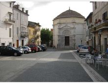 Piazza lastricata in un centro storico, con edifici residenziali ai lati e auto parcheggiate lungo la strada. Sullo sfondo si trova una chiesa in pietra con facciata decorata e grande portale, coperta da una cupola ottagonale con tetto in tegole.