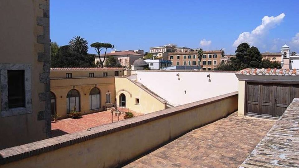 Cortile e terrazza in un complesso di edifici storici con muri intonacati gialli e bianchi, pavimentazione in mattoni e tetti in coppi. Sullo sfondo si vedono palme e altri alberi, un piccolo campanile e palazzi cittadini sotto un cielo sereno.