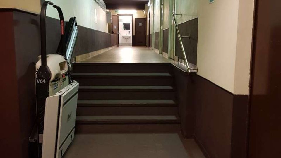 Corridoio interno con una breve rampa di scale e corrimano, illuminato da luci a parete e con soffitto in legno scuro. In alto è visibile la scritta “TOILETTE” sopra l’accesso, con porte lungo il lato destro e un dislivello verso l’area dei bagni in fondo.