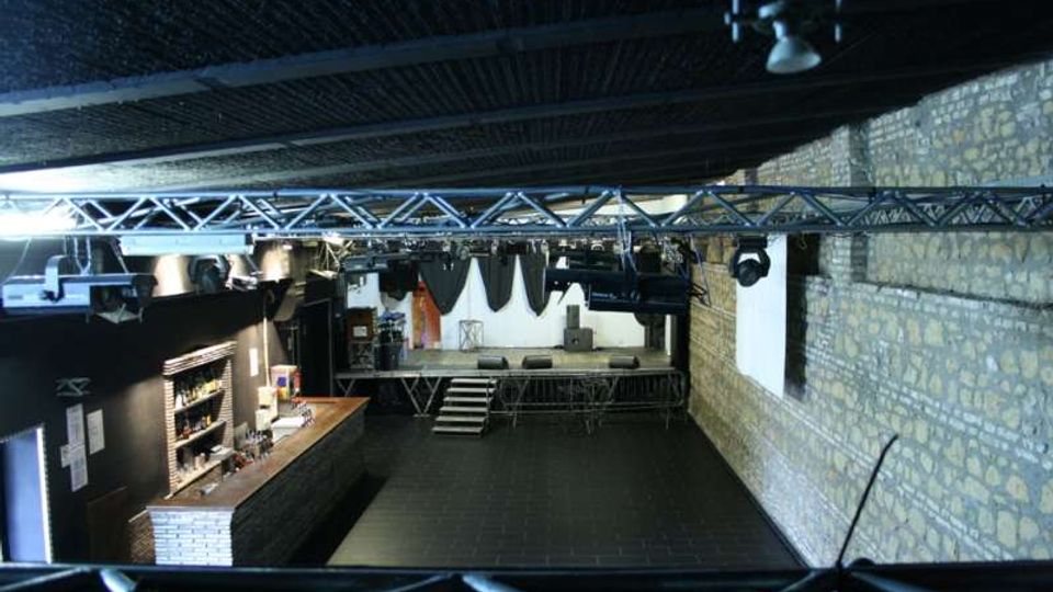 Interno di una sala eventi con pareti in pietra e soffitto scuro, vista dall’alto su un’area centrale vuota. Sul fondo c’è un palco con scalette e impianto luci su tralicci metallici, mentre a sinistra si trova un bancone bar con scaffali.