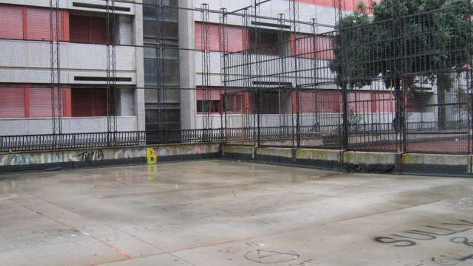 Cortile in cemento con scritte e graffiti a terra, circondato da alte recinzioni metalliche. Sullo sfondo si vede un grande edificio residenziale a più piani con balconi e persiane rosse, e un albero vicino alla rete.