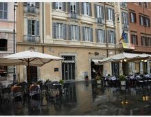 Piazza cittadina con pavimentazione bagnata dalla pioggia e tavolini all’aperto di bar sotto grandi ombrelloni e tende. Sullo sfondo, facciate di palazzi storici a più piani con persiane, balconi in ferro battuto e lampioni stradali.