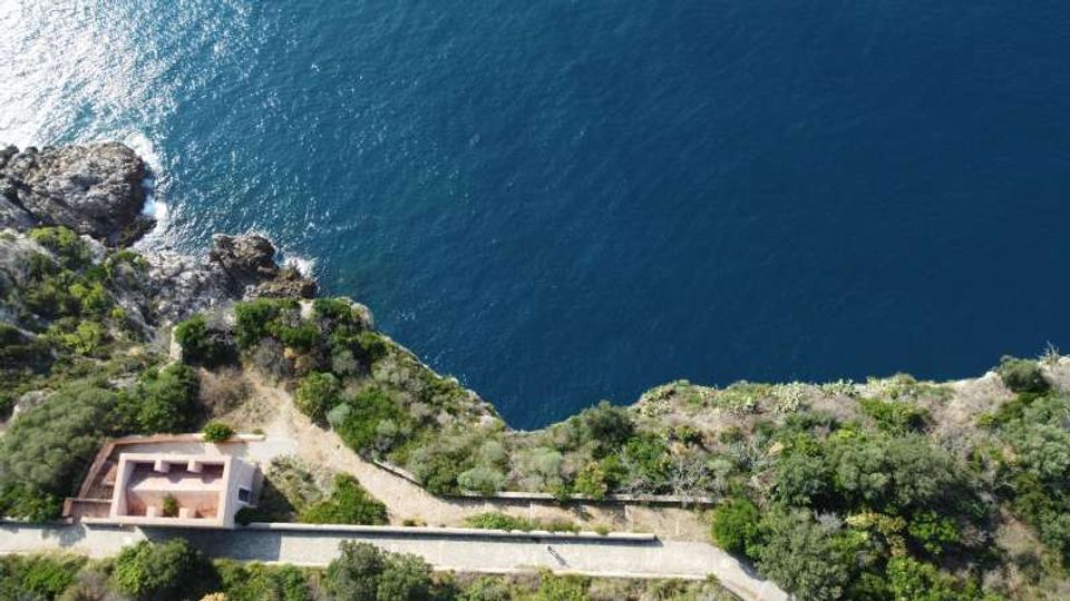Veduta aerea di una costa rocciosa con vegetazione mediterranea e mare blu intenso. In basso si vede una strada costiera e un piccolo edificio rettangolare vicino al bordo della scogliera.