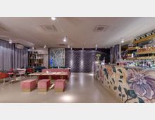 Interno di un lounge bar moderno con bancone sulla destra decorato con grandi motivi floreali e bottiglie esposte su mensole illuminate. A sinistra ci sono tavolini con panche imbottite e sedute, mentre sul fondo una parete scura con texture ondulata e tende grigie incornicia l’ambiente.