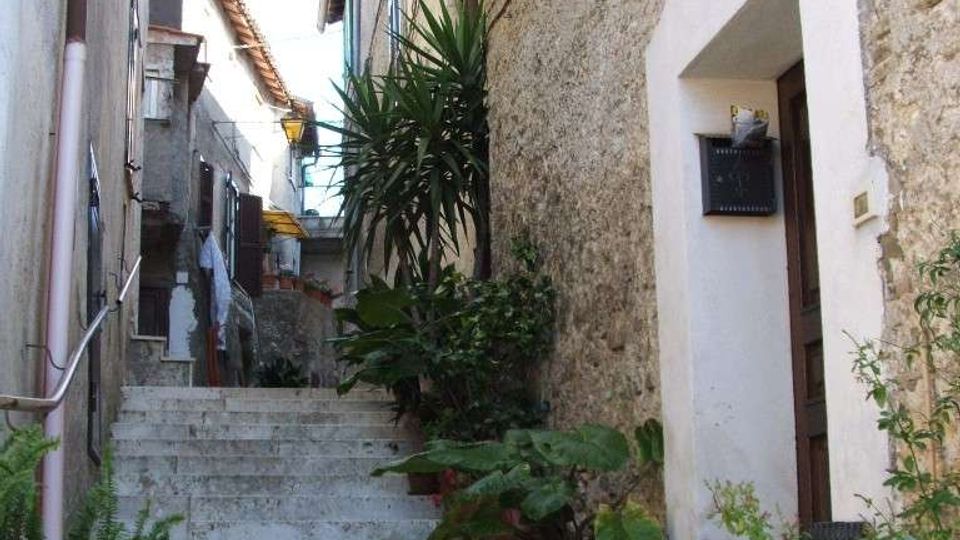 Vicolo stretto in un borgo con lunga scalinata in pietra che sale tra edifici addossati, muri in muratura e una porta incorniciata di bianco. Lungo i gradini ci sono vasi con piante verdi e lampioni a parete sospesi sopra il passaggio.
