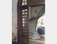 Ingresso interno di un edificio con grande porta ad arco in legno e vetri, che si apre su una scalinata in marmo curvilinea con ringhiera in ferro battuto. Sulle pareti bianche con fascia rossa sono appesi quadri e cornici, con una piccola area salotto con poltrone scure.