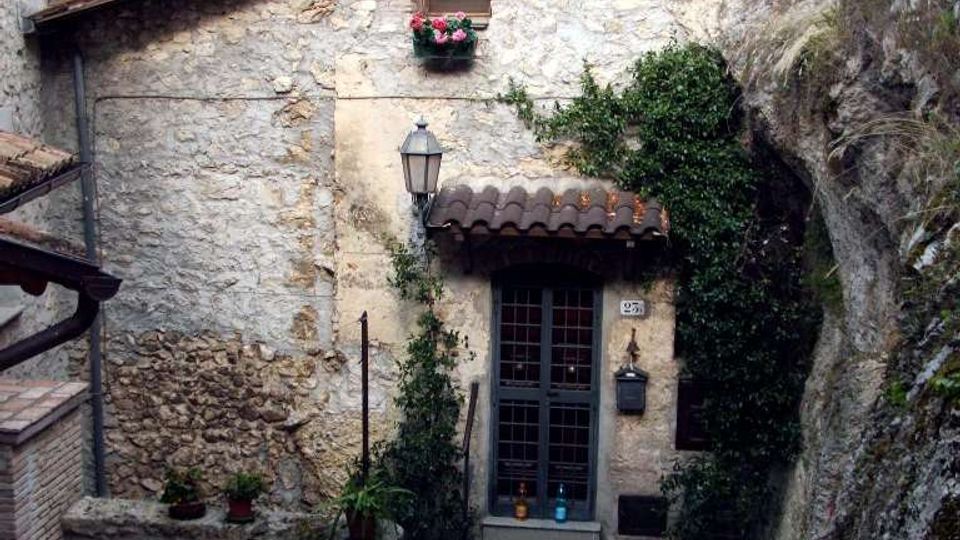 Casa in pietra addossata alla roccia lungo una stretta scalinata in selciato, con porta a griglia sotto una piccola tettoia in coppi, lanterna e piante rampicanti. Sullo sfondo si vedono montagne boscose e pareti rocciose.