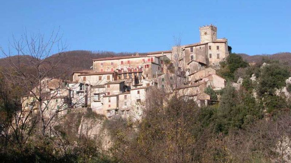 Borgo storico arroccato su un pendio, con case in pietra addossate tra loro e una torre quadrata in cima al complesso. Intorno si vedono colline boscate e vegetazione in primo piano sotto un cielo limpido.