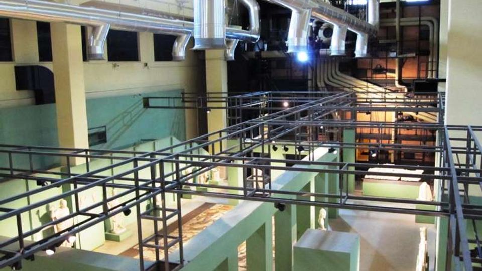 Interno di un grande edificio industriale riconvertito, con passerelle e ringhiere metalliche sopraelevate e una fitta rete di condotti e tubazioni d’aerazione a soffitto. Lo spazio è diviso da pareti verde chiaro e pilastri, con aree espositive illuminate al livello inferiore.