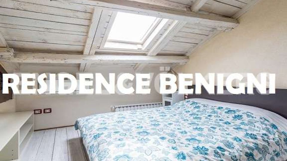 Camera da letto mansardata con soffitto in legno bianco a travi e finestra lucernario sul tetto; letto matrimoniale con copriletto a motivi floreali e pavimento in assi chiare.