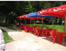 Area ristoro all’aperto in un parco, con tavoli e sedie di plastica rossi disposti lungo un vialetto curvo. Una fila di grandi ombrelloni rossi e blu con loghi Coca‑Cola copre i posti a sedere, circondati da prato e alberi.