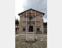 Facciata frontale di una piccola chiesa in pietra con tetto a capanna e ingresso centrale, con finestre laterali e un oculo sopra il portale. Davanti all’edificio si trova una grande croce in ferro su un basamento in muratura, in un cortile con pavimentazione in pietra e prato sotto un cielo nuvoloso.