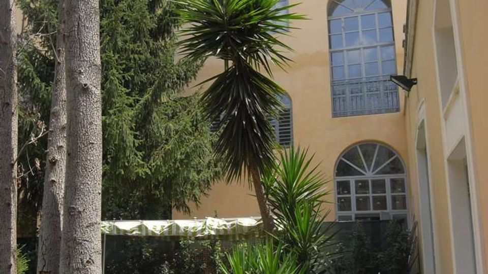 Cortile interno con ghiaia e vialetto in pietra, circondato da un edificio color crema con grandi finestre ad arco su più piani. Nello spazio verde sono presenti alberi alti e piante tropicali, con una zona ombreggiata da una copertura.