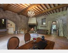Ampio soggiorno rustico con pareti in pietra, soffitto con travi in legno a vista e lampadario in cristallo. Al centro c’è un grande camino acceso, con divani e tavolino su tappeto, e un passaggio ad arco con tende che conduce a un’altra stanza.