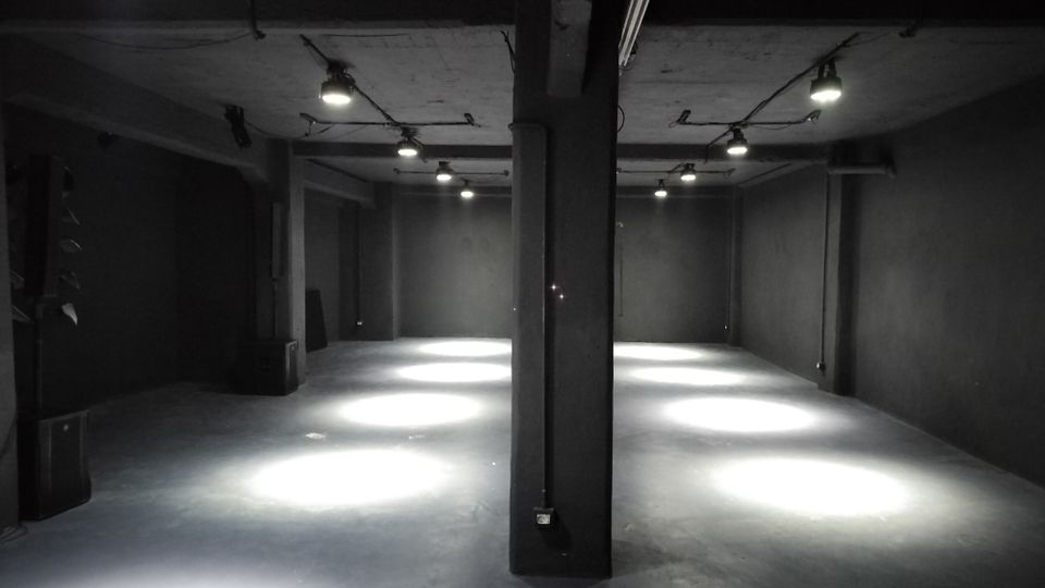 Ampio spazio interno tipo magazzino o sala industriale vuota, con pareti scure, pavimento in cemento e colonne centrali che sostengono il soffitto basso. Lampade a sospensione con cavi a vista illuminano a chiazze il pavimento lungo il corridoio centrale.