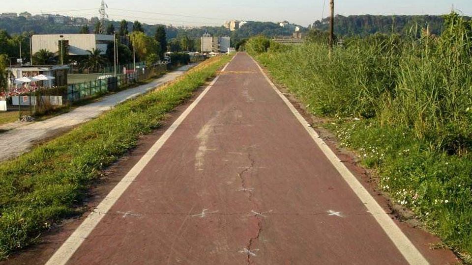 Pista ciclabile rettilinea in asfalto rosso con bordi bianchi, affiancata da erba alta e campi, che si estende verso l’orizzonte. Sullo sfondo si vedono edifici bassi, colline alberate e linee elettriche lungo il percorso.