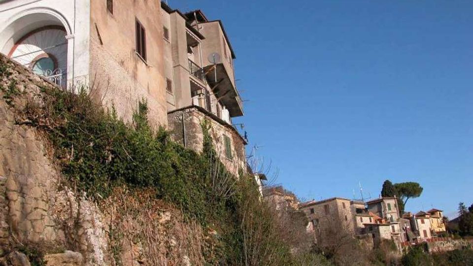 Case in pietra e intonaco costruite sul bordo di una scarpata rocciosa, con muri di contenimento e vegetazione spontanea sul pendio. Sullo sfondo si vede un piccolo borgo collinare con altri edifici, un grande albero e cielo sereno.