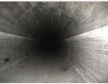 Interno di un tunnel sotterraneo in cemento con volta ad arco e pareti scanalate, che si estende in profondità verso un’area buia. Il pavimento è in cemento e presenta crepe e segni di usura lungo i bordi.
