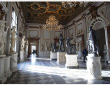 Ampia sala museale in stile neoclassico con pavimento a scacchi in marmo, grandi finestre laterali e soffitto a cassettoni decorato con lampadari. Numerose statue su piedistalli e nicchie lungo le pareti ornano lo spazio, con un passaggio in fondo alla sala.