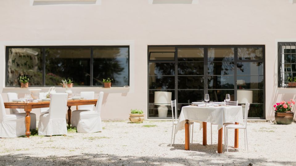 Cortile esterno con tavoli apparecchiati e sedie bianche su ghiaia, davanti alla facciata chiara di un edificio con grandi finestre e porte vetrate. Vasi di piante e fiori decorano l’area vicino alle aperture.