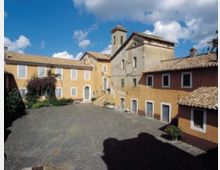 Cortile in pietra di un complesso storico con edifici intonacati color ocra e rosso, finestre con cornici bianche e tetti in coppi. Sullo sfondo si vede una chiesa con campanile e facciate in muratura sotto un cielo azzurro con nuvole.