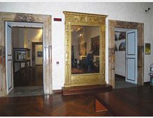 Interno di museo con pavimento in parquet scuro e pareti bianche, con due porte aperte incorniciate in marmo che conducono a sale espositive. Al centro è presente un grande specchio con cornice dorata decorata, su pedana in legno, e sulle pareti si intravedono dipinti e teche.