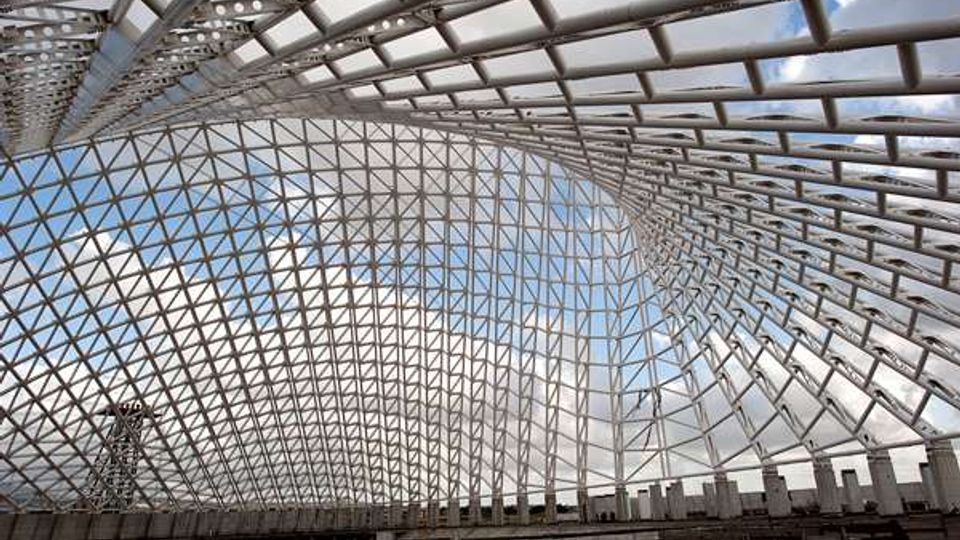 Interno di un grande atrio coperto da una struttura reticolare in acciaio con ampie superfici vetrate, che forma una volta curva e lascia vedere il cielo. In basso si notano gradinate e una fascia di parapetti lungo il perimetro.