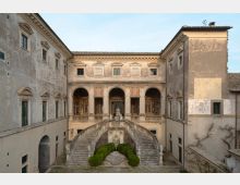 Cortile interno di un palazzo storico con facciate intonacate e finestre incorniciate, affacciato su una loggia ad archi. Al centro si trova una scenografica scalinata doppia in pietra con balaustre e statue, che scende verso un piccolo giardino geometrico con siepi e fontana.