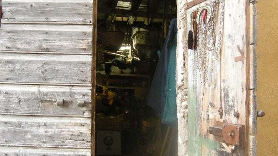 Ingresso di un vecchio capanno o magazzino con grandi porte in legno grezzo aperte e un arco con finestra in vetro sopra. All’interno si intravedono attrezzi e materiali accatastati in un ambiente buio e polveroso.