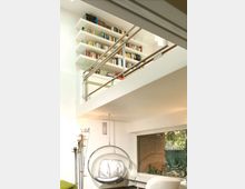 Soggiorno moderno a doppia altezza con soppalco e ringhiera metallica, pareti bianche e grandi scaffali pieni di libri. In basso si vede una poltrona sospesa trasparente e una grande finestra con vista su un piccolo giardino e un muro in mattoni.