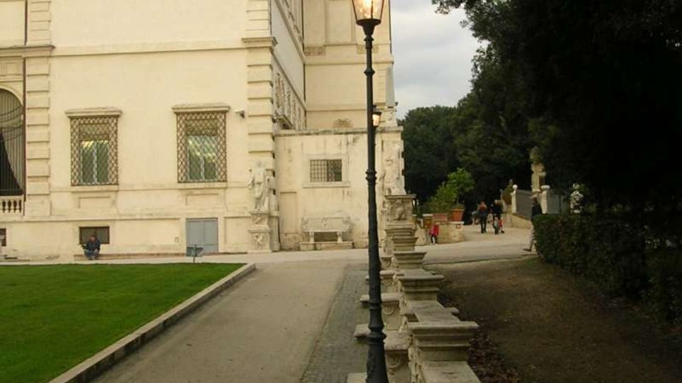Viale pedonale accanto a un grande edificio storico in pietra chiara, con finestre alte e dettagli decorativi sulla facciata. Lungo il percorso ci sono un lampione in stile classico e una balaustra in pietra, con alberi fitti e area verde sul lato opposto.