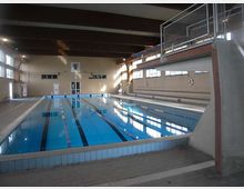 Piscina coperta con vasca lunga a corsie delimitate da galleggianti, acqua azzurra e bordo piastrellato. L’ambiente è un grande locale con travi a vista sul soffitto, ampie finestre laterali e una passerella sopraelevata con ringhiera metallica su un lato.
