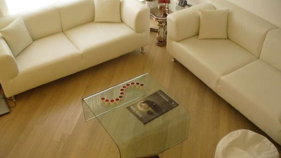 Soggiorno luminoso con pavimento in legno chiaro, due divani bianchi disposti ad angolo e un tavolino basso in vetro curvato al centro. Sullo sfondo c’è un tavolino laterale con lampada rossa e piccoli oggetti decorativi.