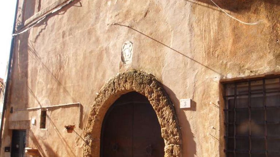 Facciata di un edificio antico in intonaco color ocra, con grande portale ad arco in pietra e portone in legno. Si vedono finestre con grate, cavi esterni lungo il muro e una strada lastricata in pietra con vasi di fiori.