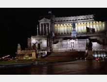Veduta notturna del monumento del Vittoriano a Roma, con la lunga facciata a colonne e le terrazze illuminate. In primo piano si vedono la scalinata monumentale e la statua equestre centrale, con strada e luci urbane ai margini.