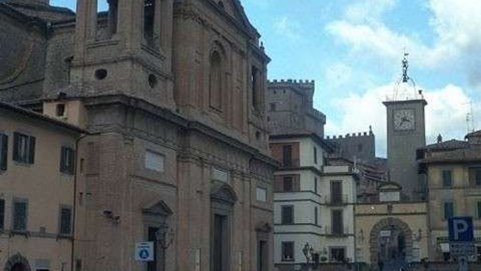 Veduta di una piazza in un centro storico con una grande chiesa in pietra sulla sinistra e edifici storici lungo la strada. Sullo sfondo si vedono una torre con orologio e un arco di passaggio che conduce a una via più stretta.