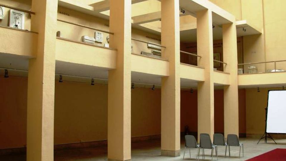 Cortile interno di un edificio con grandi colonne e ballatoi su più livelli, con pareti color beige e ringhiere metalliche. Nello spazio a terra ci sono alcune sedie, un tappeto rosso e uno schermo di proiezione su un lato.