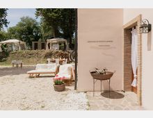 Cortile esterno di una struttura rustica con parete in intonaco chiaro e porta incorniciata in mattoni, illuminata da una lanterna. Davanti ci sono lettini e tavoli su ghiaia, una catasta di legna e gazebo bianchi tra alberi sullo sfondo.