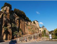 Strada ai piedi di una parete rocciosa scoscesa con vegetazione, sormontata da resti di mura in pietra e da un edificio storico. In basso si vede un parcheggio con segnaletica e un piccolo arco d’ingresso lungo il percorso.