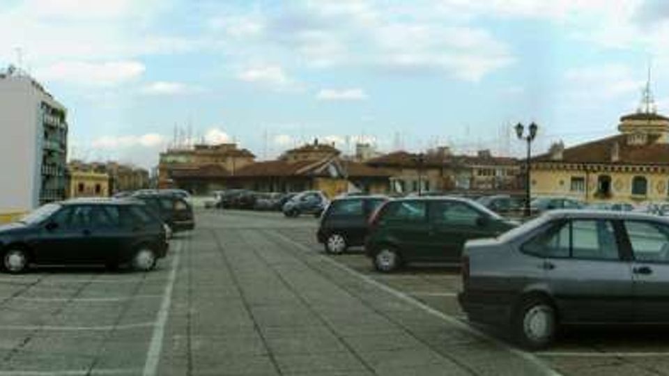 Ampio parcheggio sopraelevato con pavimentazione in cemento e auto parcheggiate in file, affiancato da una ringhiera gialla e un muretto. Sullo sfondo si vede un panorama urbano con edifici residenziali e strutture storiche a tetto basso sotto un cielo parzialmente nuvoloso.