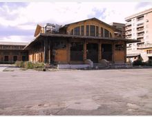 Grande edificio industriale in mattoni, con ampie finestre ad arco e una tettoia sporgente sostenuta da colonne, affacciato su un vasto piazzale asfaltato. Sullo sfondo si vedono altre strutture del complesso e palazzi residenziali moderni.