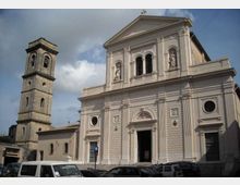 Facciata di una chiesa in stile neoclassico con frontone triangolare, colonne e tre archi superiori con statue in nicchie; a sinistra si vede un campanile in pietra a torre. Davanti all’edificio c’è una piazza con auto parcheggiate sotto un cielo parzialmente nuvoloso.