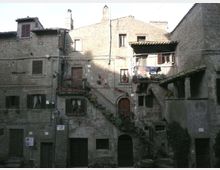 Facciata di un complesso di case in pietra in un borgo storico, con finestre e portoni scuri, balconi con vasi e una scalinata esterna in muratura con ringhiera. Un piccolo loggiato coperto e tetti in coppi completano l’insieme delle costruzioni addossate tra loro.