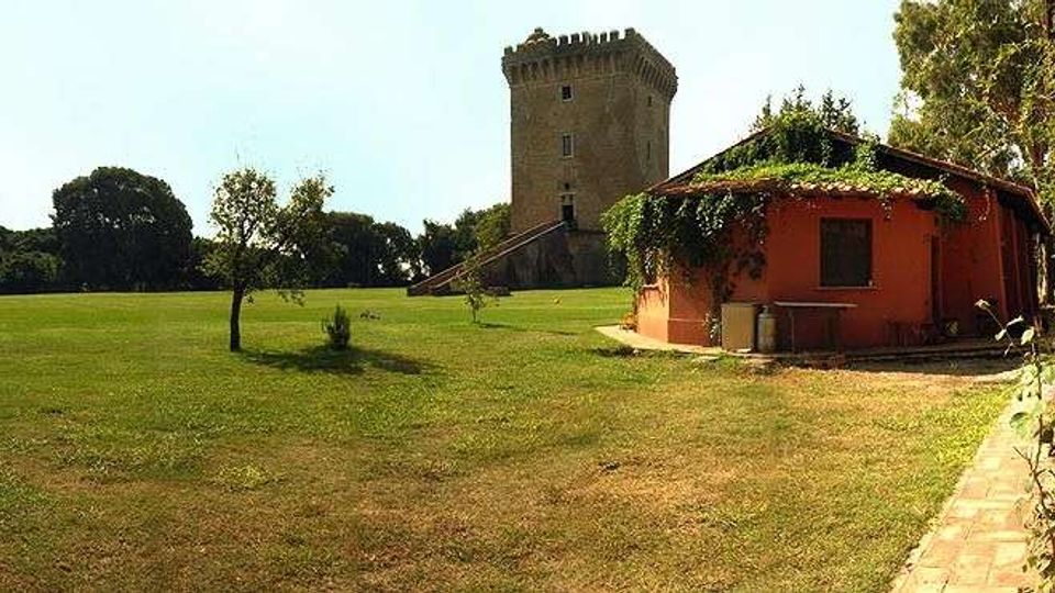 Ampio prato con alberi sparsi e un sentiero in terra battuta, con una piccola costruzione bassa color arancione parzialmente coperta da vegetazione. Sullo sfondo si vede una torre in pietra di tipo medievale con merlature, circondata da alberi.