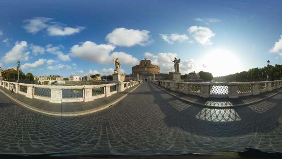 Veduta panoramica di un ponte in pietra con pavimentazione in sampietrini, balaustre e statue lungo i lati, che attraversa un fiume. Sullo sfondo si vede un grande edificio circolare fortificato e, a destra, una fila di alberi lungo la riva sotto un cielo con nuvole.